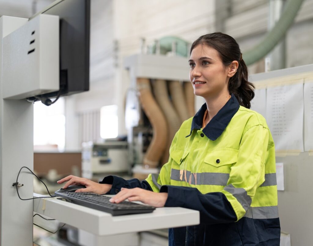 Woman, wearing a safety jacket, operates a CNC machine in an industrial setting