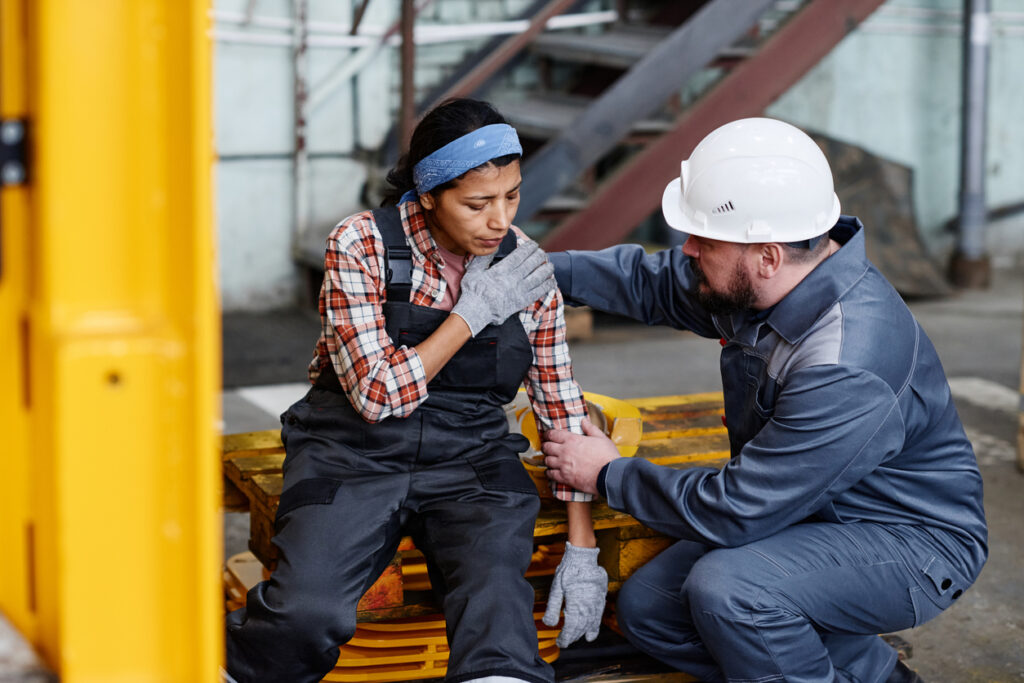 In an industrial setting, a female employee is being assisted with a shoulder injury by a male colleague.