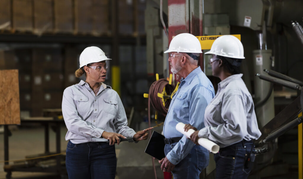 Three mature workers in safety gear converse in a metal fabrication factory. There are two women and one man.