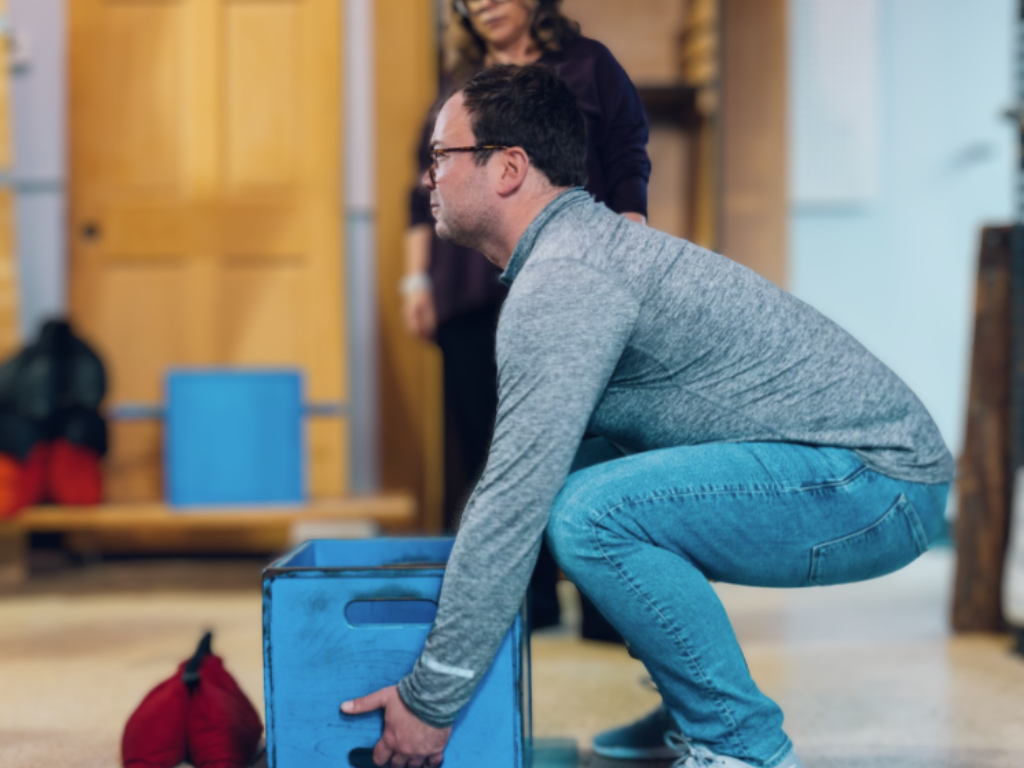 Man, using proper lifting technique, bends his legs as he prepares to life a NIOSH box during a physical agility test. The female on-site testing specialist observes from the background.