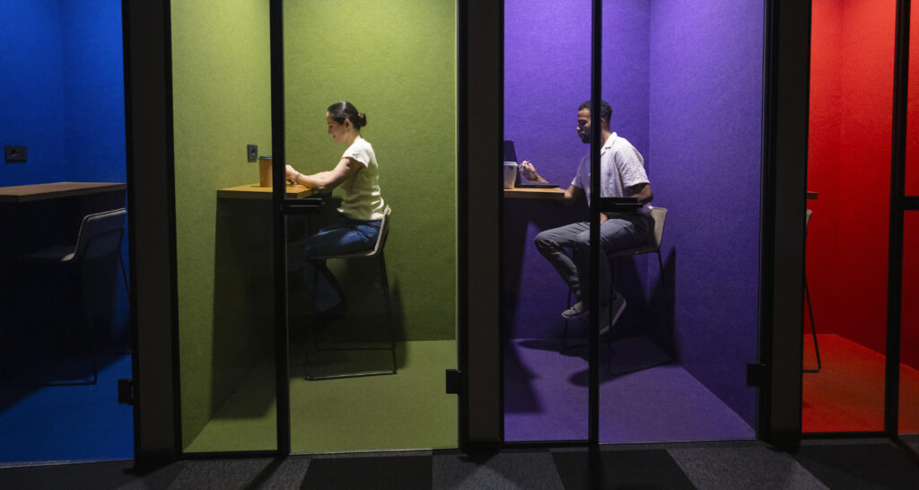 A group of business professionals of various ethnicities work diligently in vibrant, separate office cubicles, each absorbed in tasks involving phones and computers.