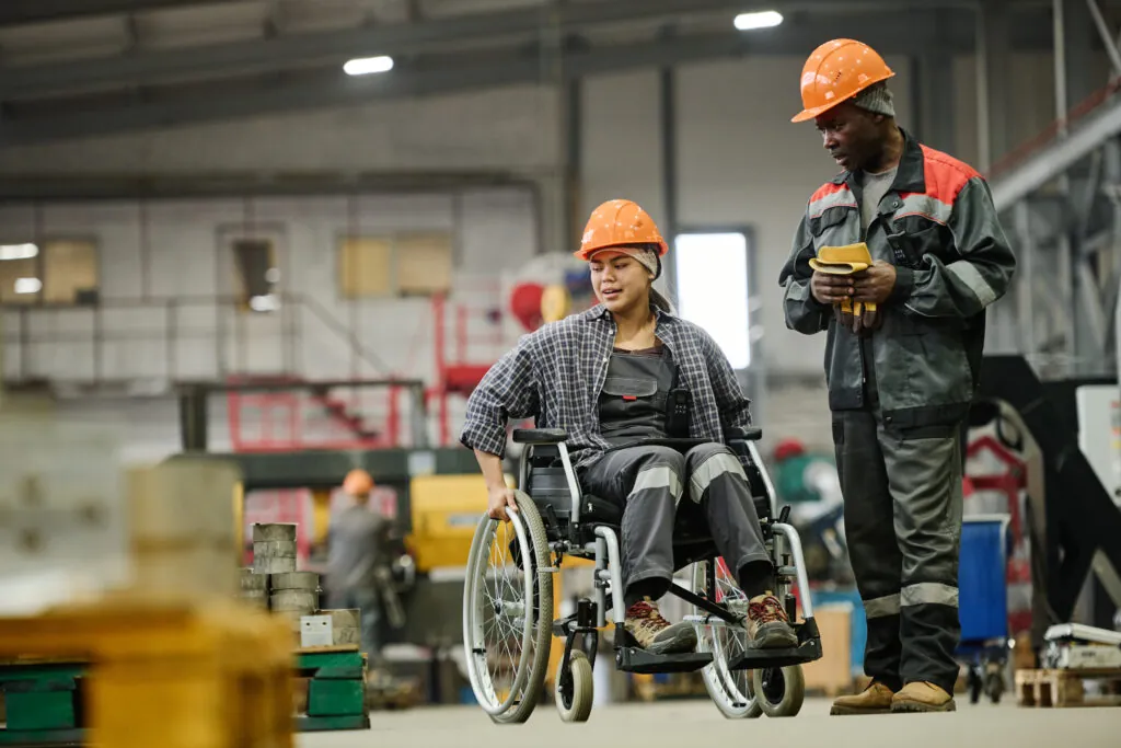 Wheelchair-using worker conversing with a colleague in an industrial warehouse setting.