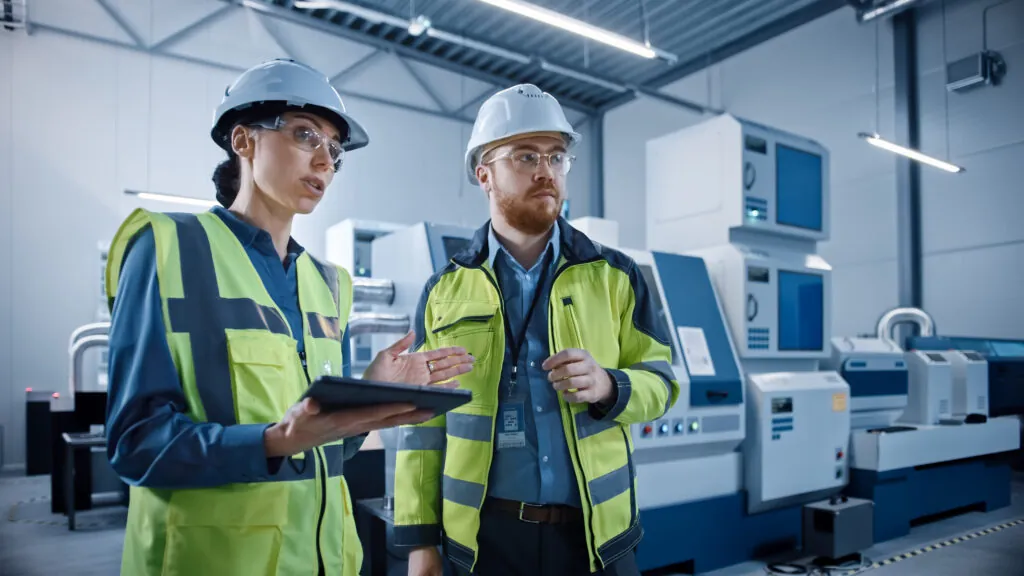 Modern Factory: Female Project Manager and Male Engineer Standing Wearing Safety Jackets, Hard Hats, Work in Workshop, Talking, Using Digital Tablet and Monitoring CNC Machinery Assembly Line