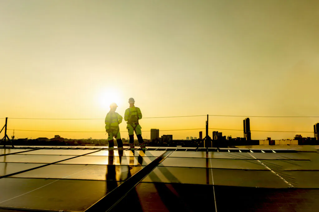 Two engineers in safety clothing with helmet check solar system at rooftop solar farm with city view. Engineers installing solar panels. Man and woman workers inspecting solar panels on roof at sunset