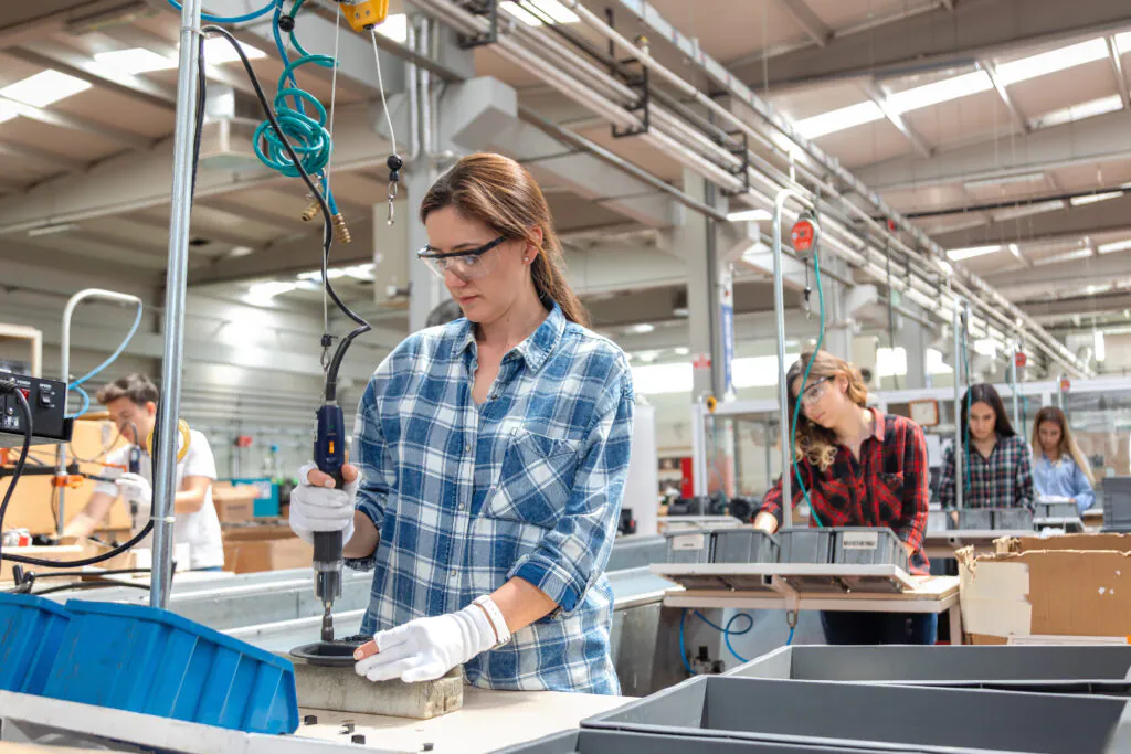 Professional factory skilled workers wearing safety glasses and protective gloves operating powered tool while assembling equipment, representing engineering industry, industrial manufacturing process, quality control, technical labor, workplace safety and modern production environment with organized assembly station